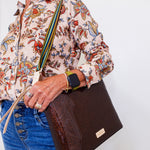 Person holding a brown clutch bag with a floral shirt and blue jeans against a white background