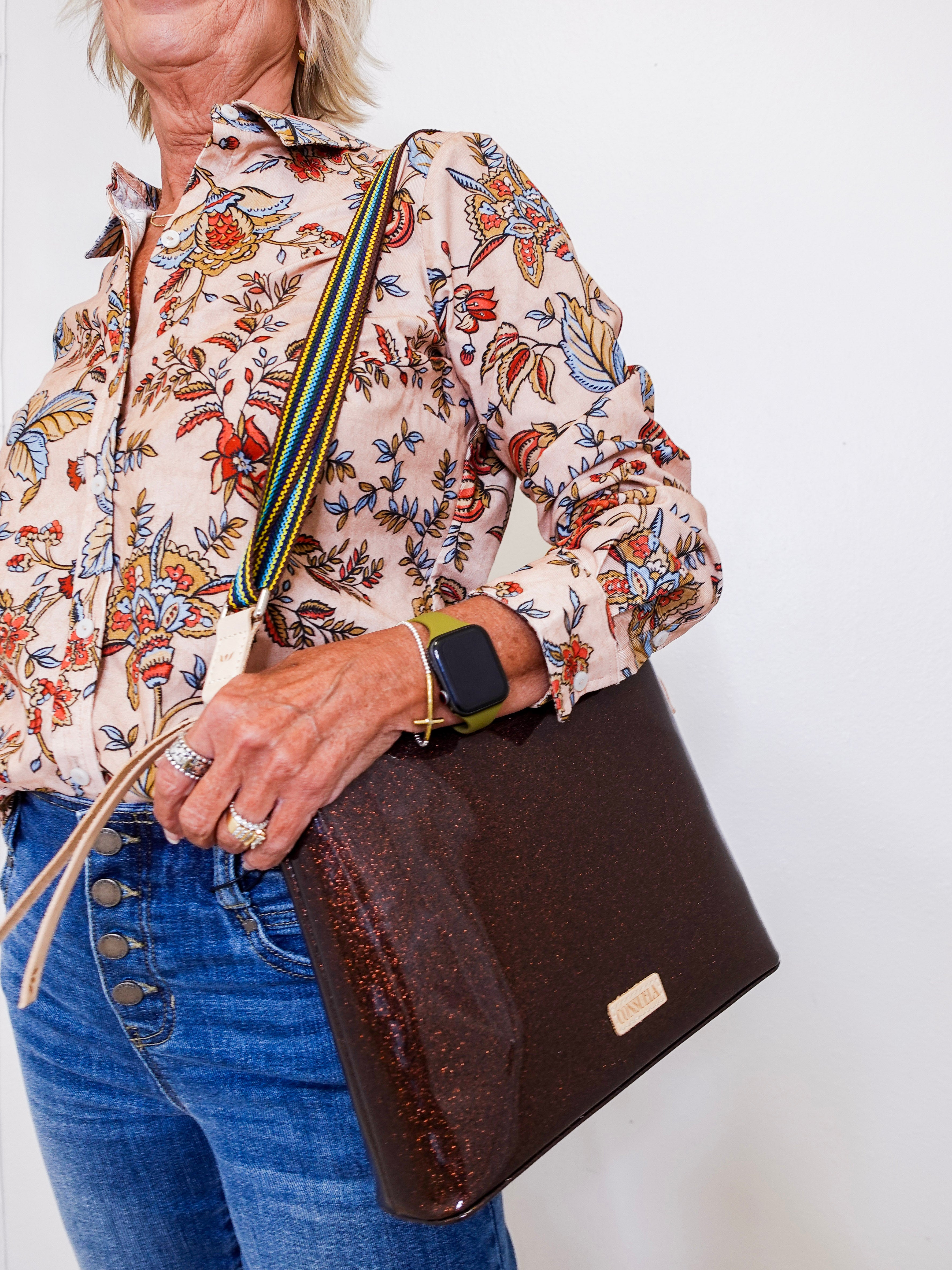 Person holding a brown clutch bag with a floral shirt and blue jeans against a white background