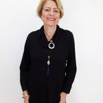 A smiling woman with short blonde hair wearing a black long-sleeve top and layered necklaces, standing against a plain white background.