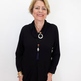 A smiling woman with short blonde hair wearing a black long-sleeve top and layered necklaces, standing against a plain white background.