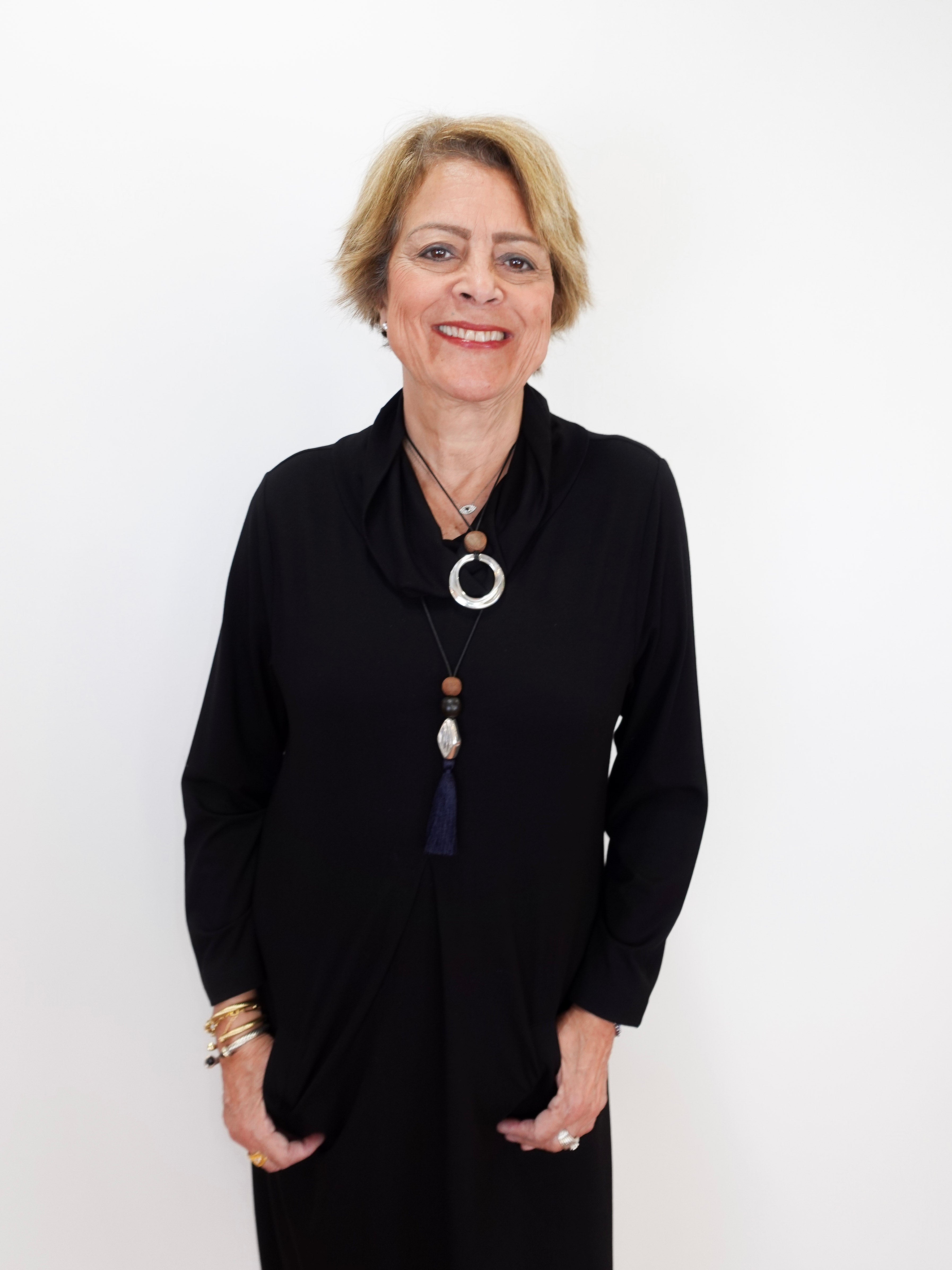 A smiling woman with short blonde hair wearing a black long-sleeve top and layered necklaces, standing against a plain white background.