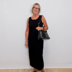 Woman in a black dress holding a black handbag against a white wall.