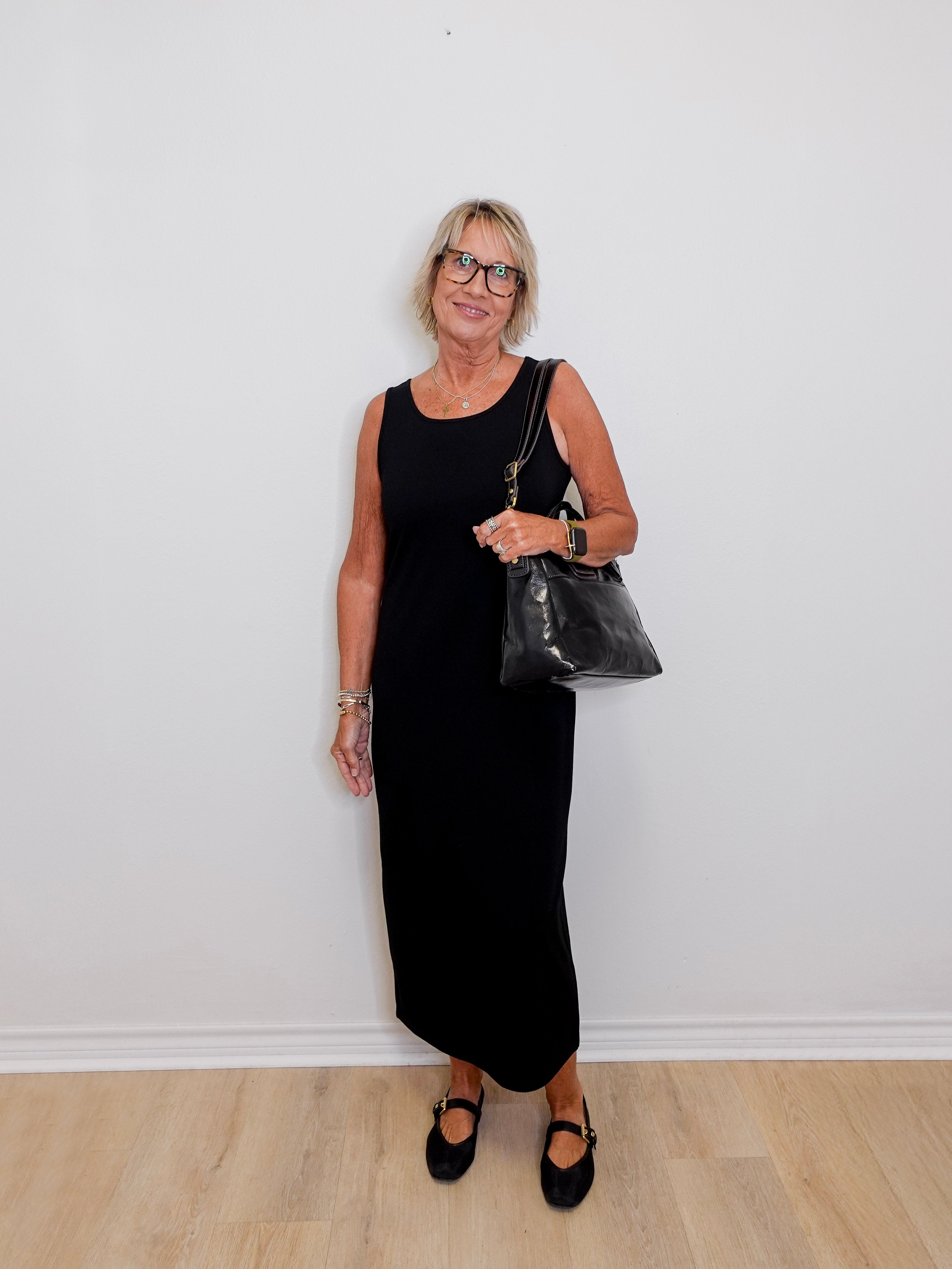 Woman in a black dress holding a black handbag against a white wall.