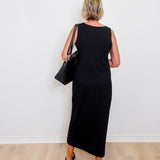 Person wearing a black sleeveless dress holding a black handbag against a white wall.