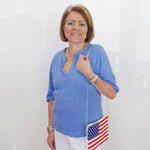 Woman wearing a blue shirt and white pants, holding an American flag bag against a white background