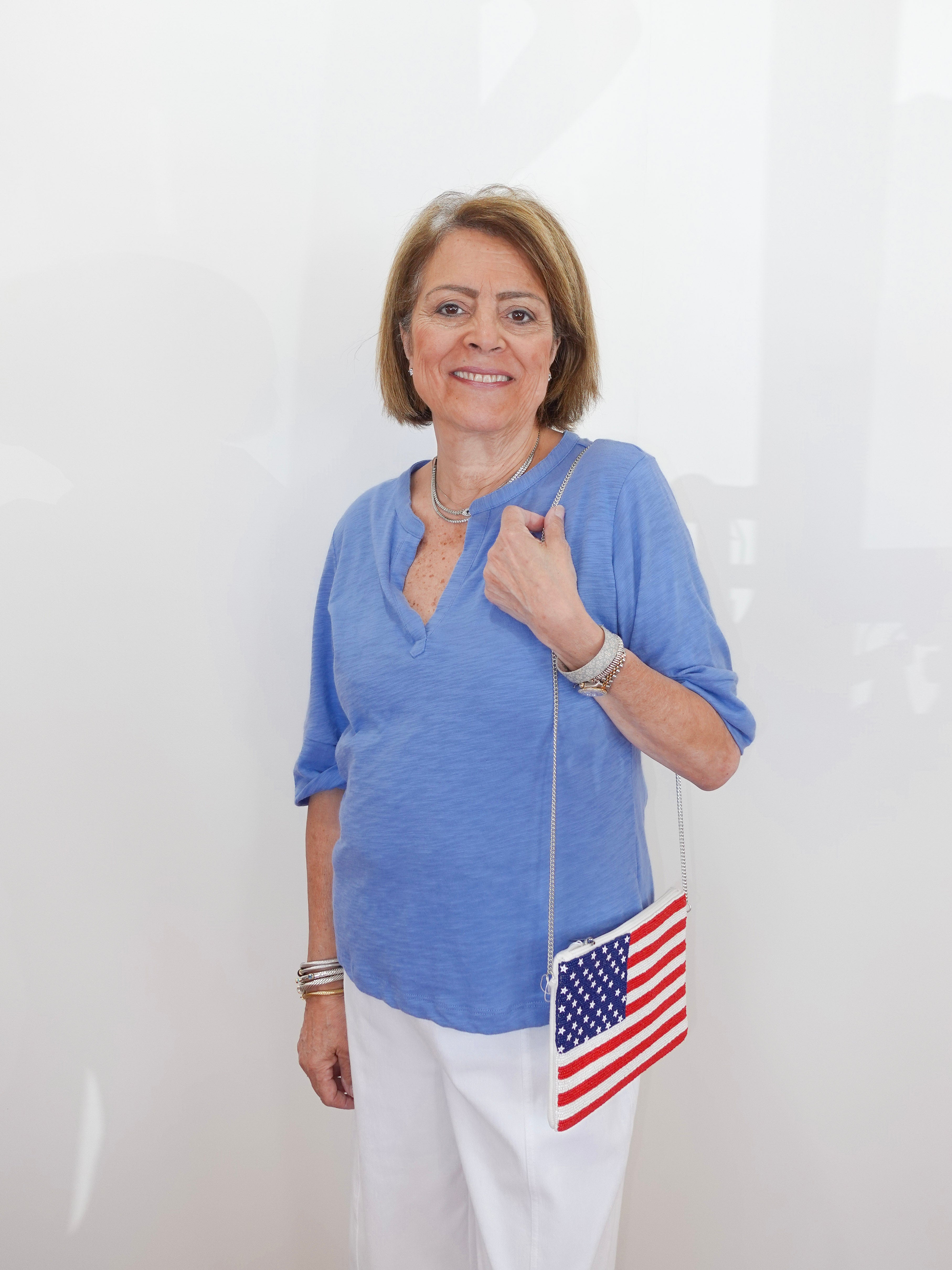 Woman wearing a blue shirt and white pants, holding an American flag bag against a white background