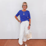 Woman wearing a blue top and white pants, holding a beige bag against a white wall.