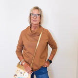 Woman modeling a tan cowl neck sweater and blue jeans in front of a white wall.