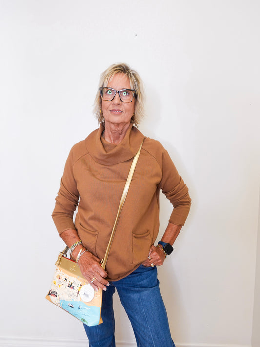 Woman modeling a tan cowl neck sweater and blue jeans in front of a white wall.