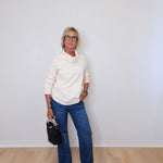 A woman wearing a cowl neck sweater in cream with blue pants against a white wall. 