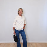 A woman wearing a cowl neck sweater in cream with blue pants against a white wall. 
