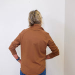 A woman wearing a tan cowl neck sweater with blue jeans against a white wall. 