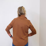 A woman wearing a tan cowl neck sweater with blue jeans against a white wall. 