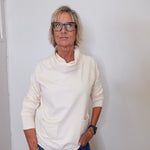 A woman wearing a cowl neck sweater in cream with blue pants against a white wall. 