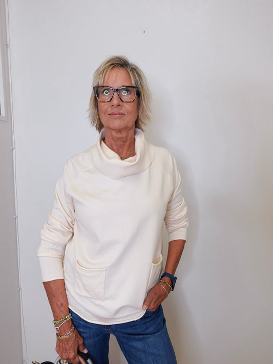 A woman wearing a cowl neck sweater in cream with blue pants against a white wall. 