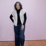 Woman wearing a white knit vest over a black top and jeans against a pink wall.