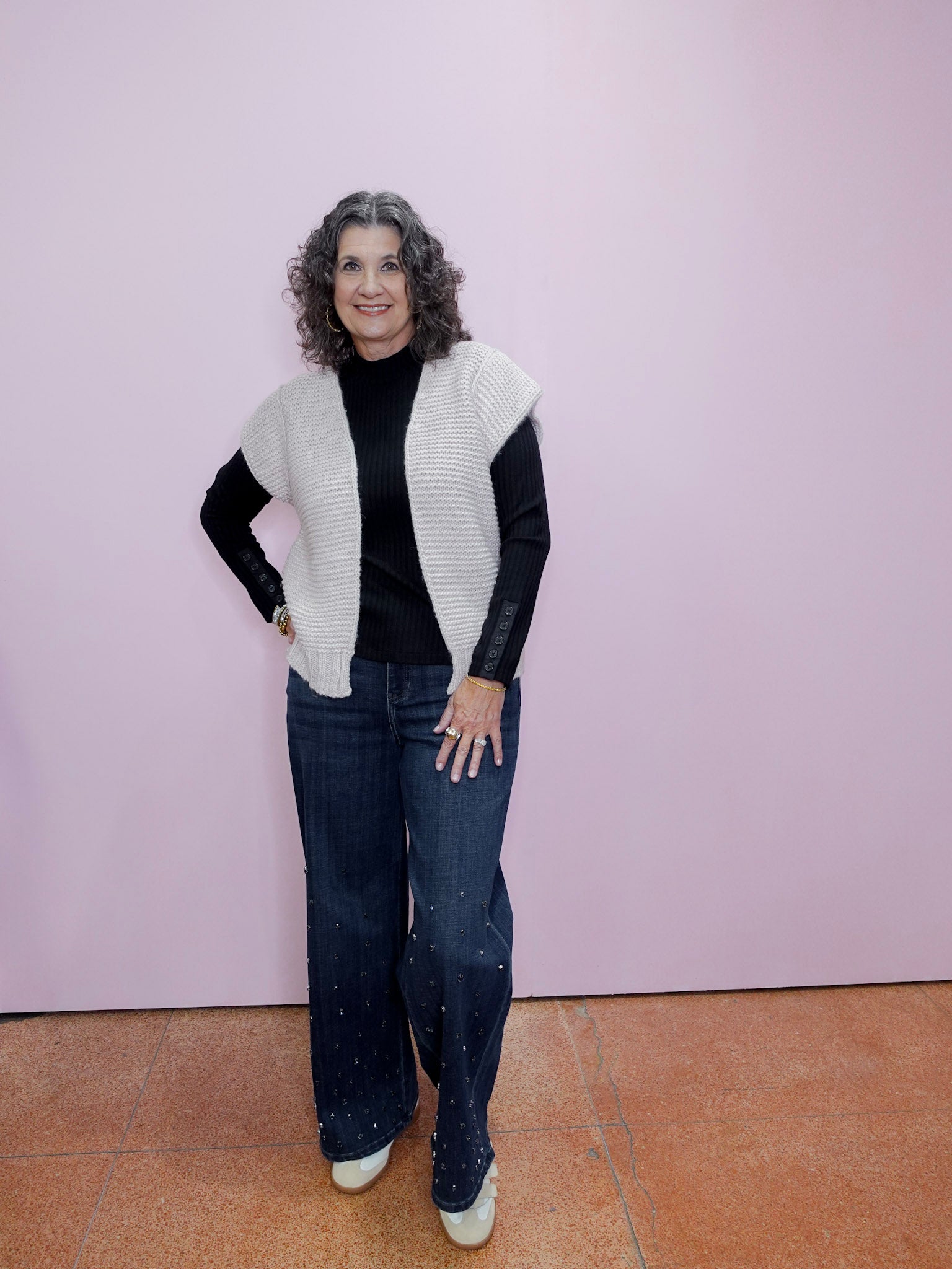 Woman wearing a white knit vest over a black top and jeans against a pink wall.