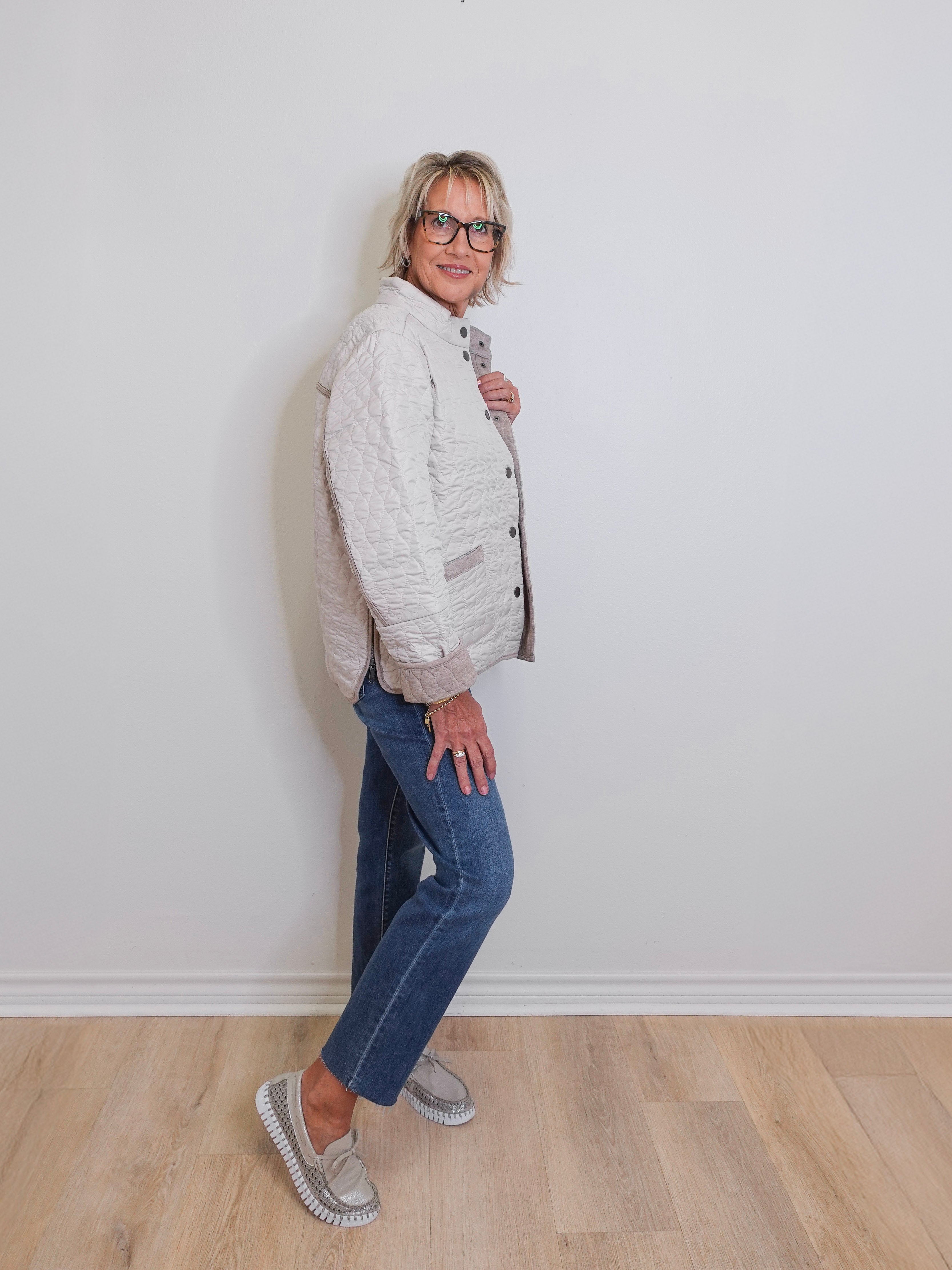 Woman wearing a white quilted jacket and blue jeans against a plain white wall.