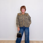 Person wearing a leopard print jacket and blue jeans, holding a black handbag against a white wall.