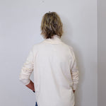 A woman wearing a cowl neck sweater in cream with blue pants against a white wall. 