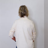 A woman wearing a cowl neck sweater in cream with blue pants against a white wall. 