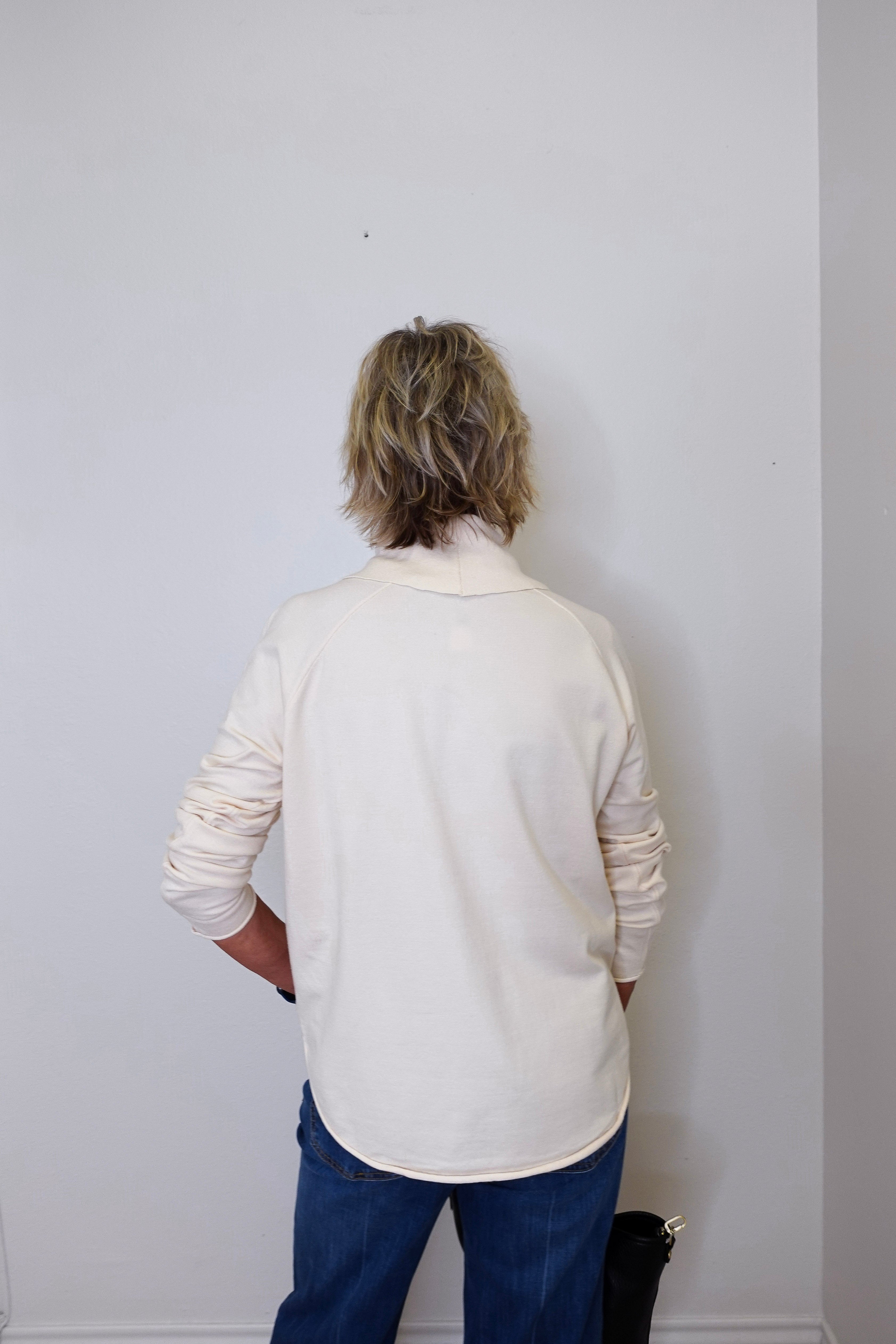 A woman wearing a cowl neck sweater in cream with blue pants against a white wall. 
