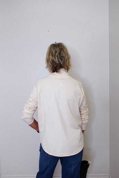 A woman wearing a cowl neck sweater in cream with blue pants against a white wall. 