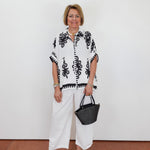 Woman wearing a black and white patterned top with white pants, holding a black handbag against a white background.