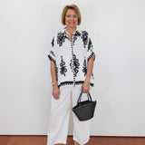 Woman wearing a black and white patterned top with white pants, holding a black handbag against a white background.