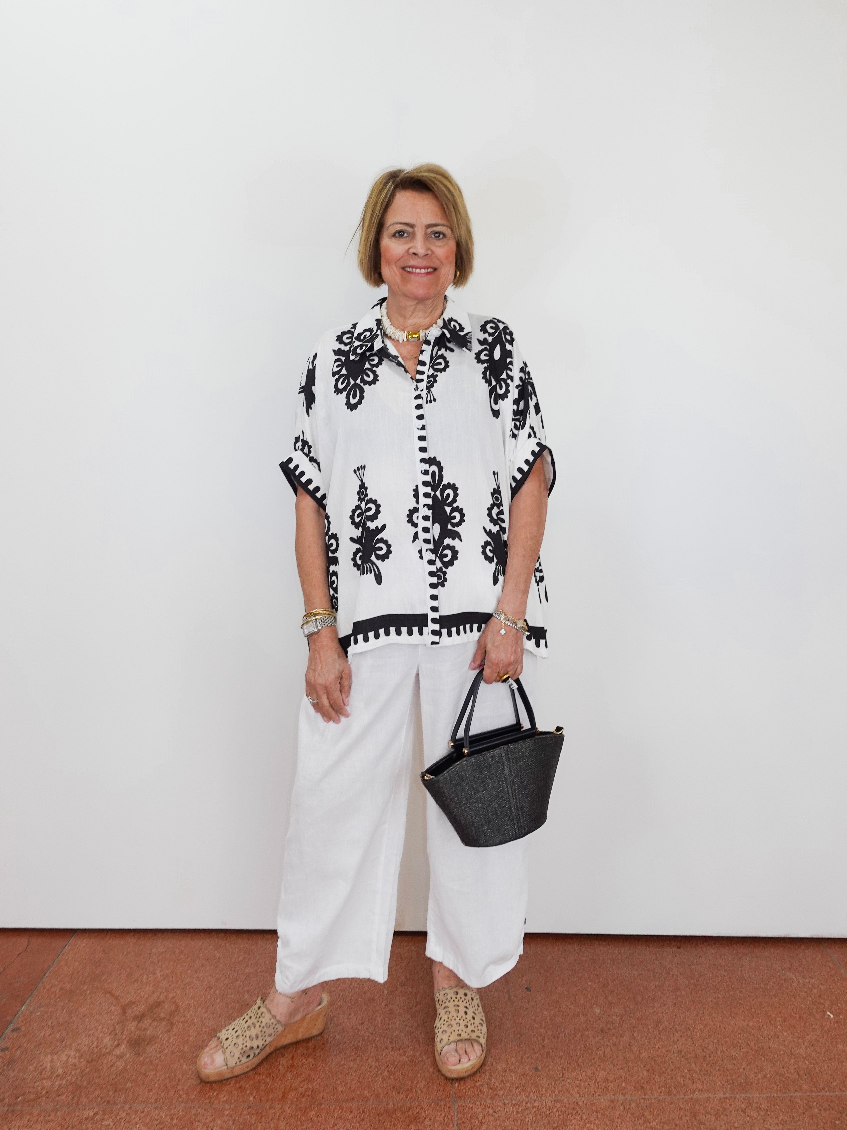 Woman wearing a black and white patterned top with white pants, holding a black handbag against a white background.