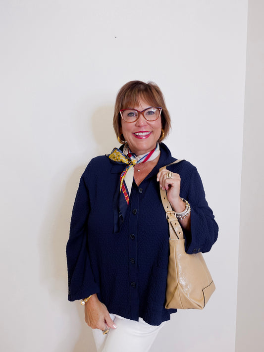 Woman wearing a navy cardigan, white pants, and holding a beige handbag against a plain background
