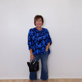 Woman wearing a blue floral blouse and jeans, holding a black handbag against a white wall.