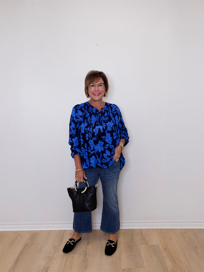 Woman wearing a blue floral blouse and jeans, holding a black handbag against a white wall.