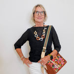 Woman wearing a black top, white pants, and glasses, holding a colorful bag against a white background