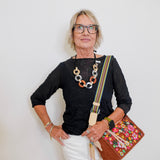 Woman wearing a black top, white pants, and glasses, holding a colorful bag against a white background