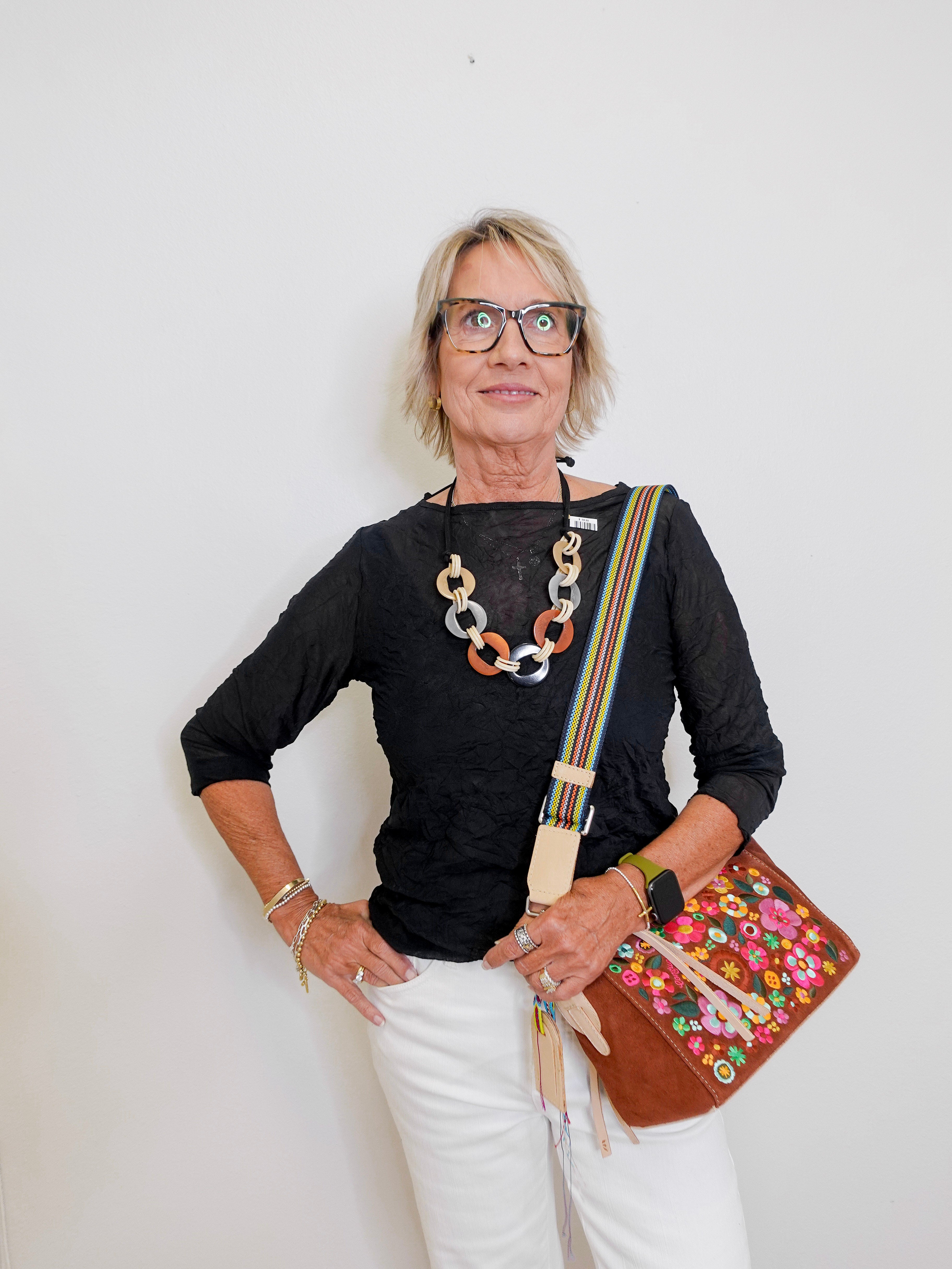 Woman wearing a black top, white pants, and glasses, holding a colorful bag against a white background