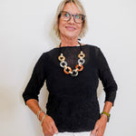 Woman wearing a black top and white pants with a colorful necklace against a plain background
