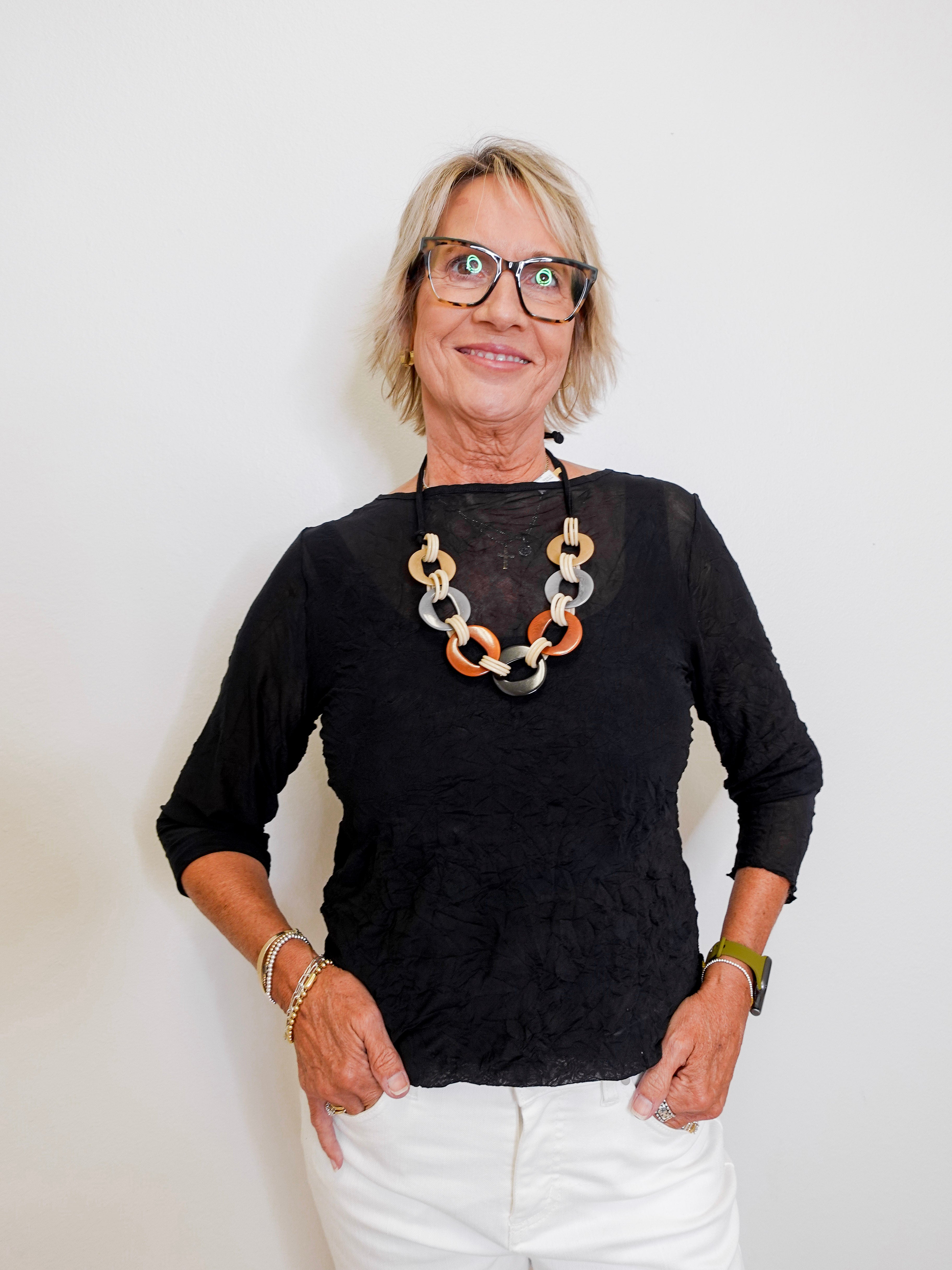 Woman wearing a black top and white pants with a colorful necklace against a plain background