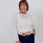 Woman wearing a white long-sleeve top and blue jeans with a gold belt, standing against a plain background.