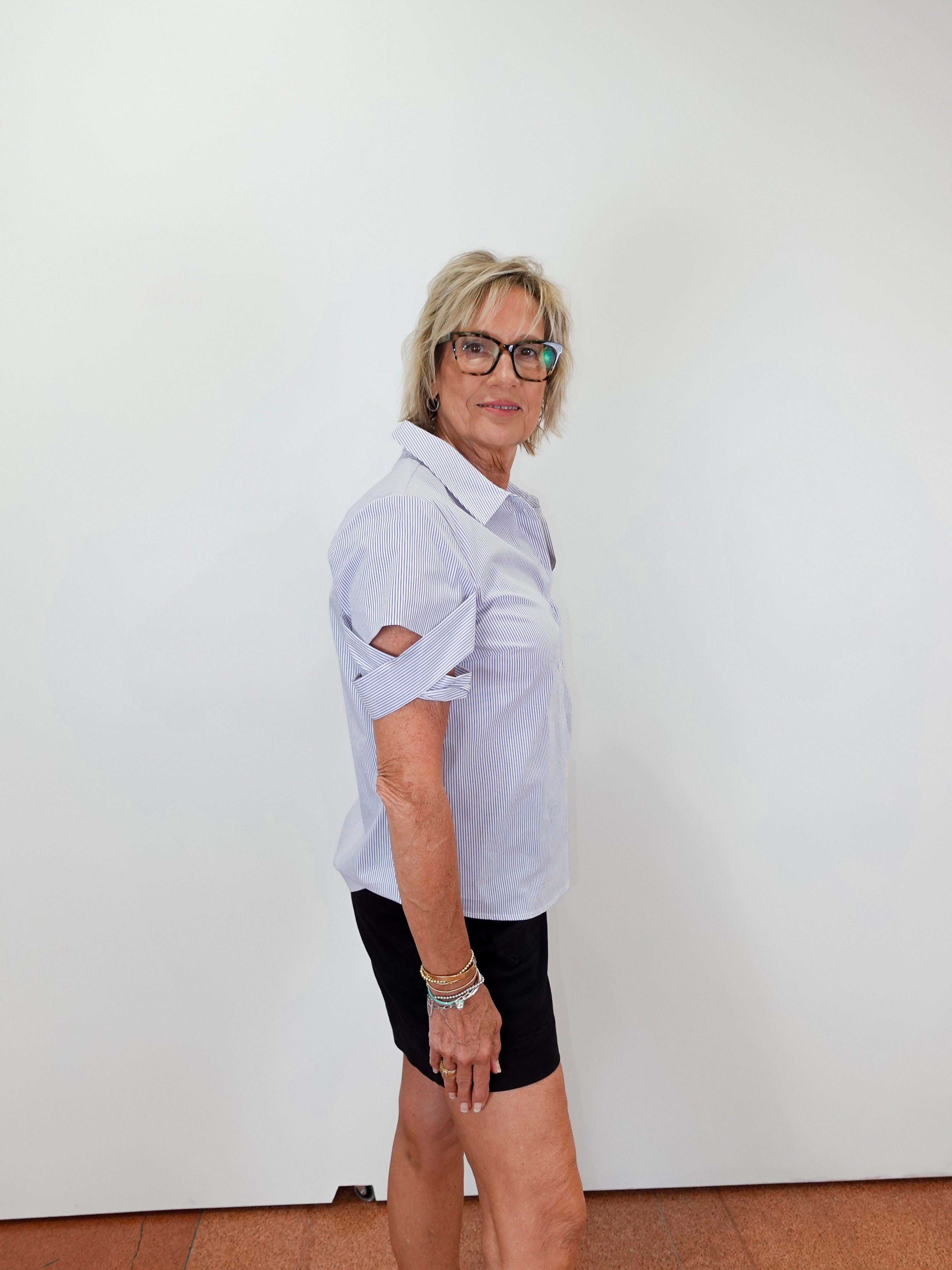 Woman wearing a light blue shirt, black shorts, and white sneakers standing against a white wall.