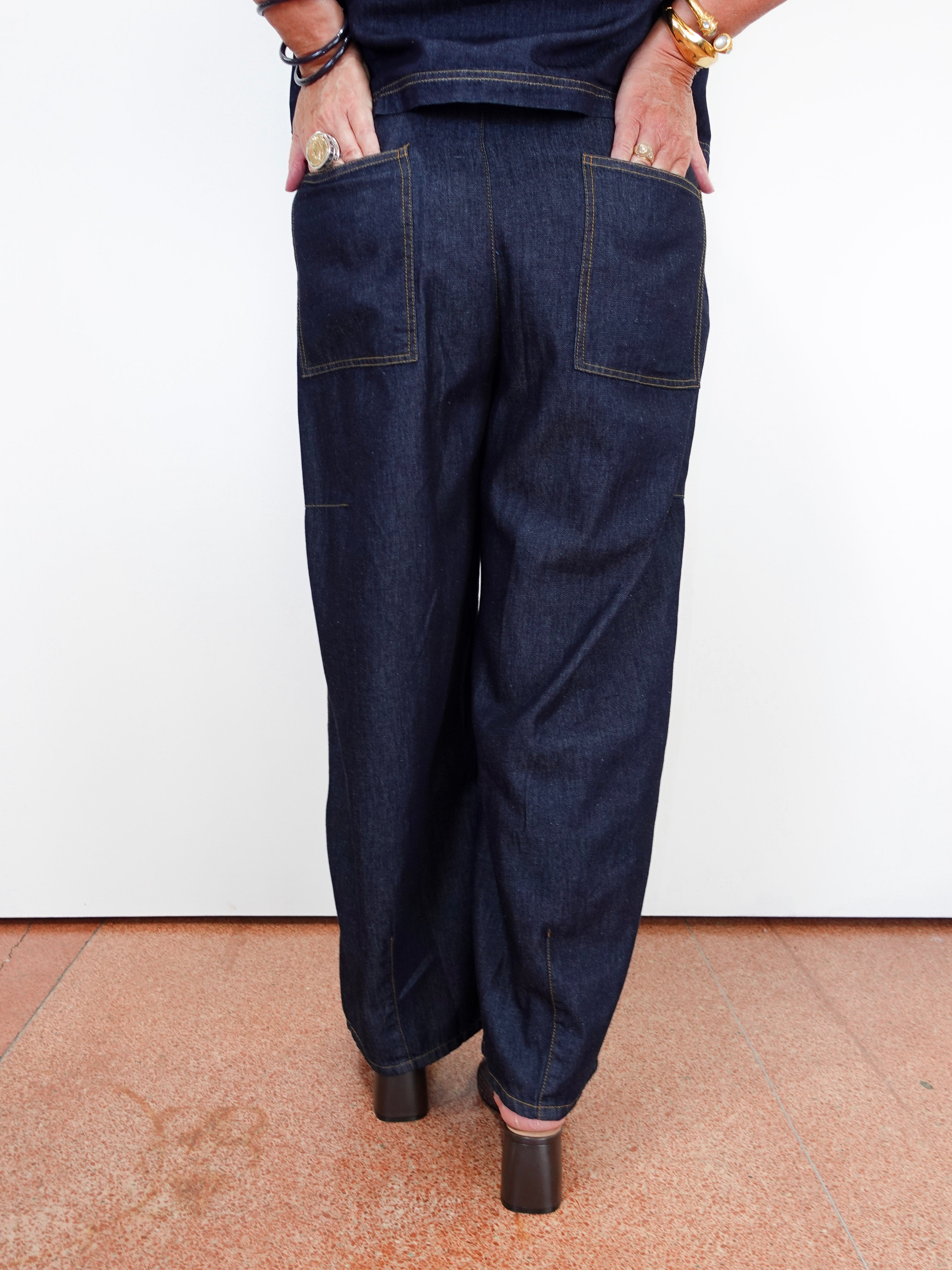 A person shown from the back wearing dark indigo wide-leg denim pants with large patch pockets, standing against a white wall on a terracotta-colored floor.
