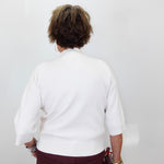 A person viewed from behind wearing a white knit sweater with three-quarter sleeves and burgundy pants, standing against a plain light gray wall.
