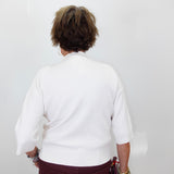 A person viewed from behind wearing a white knit sweater with three-quarter sleeves and burgundy pants, standing against a plain light gray wall.