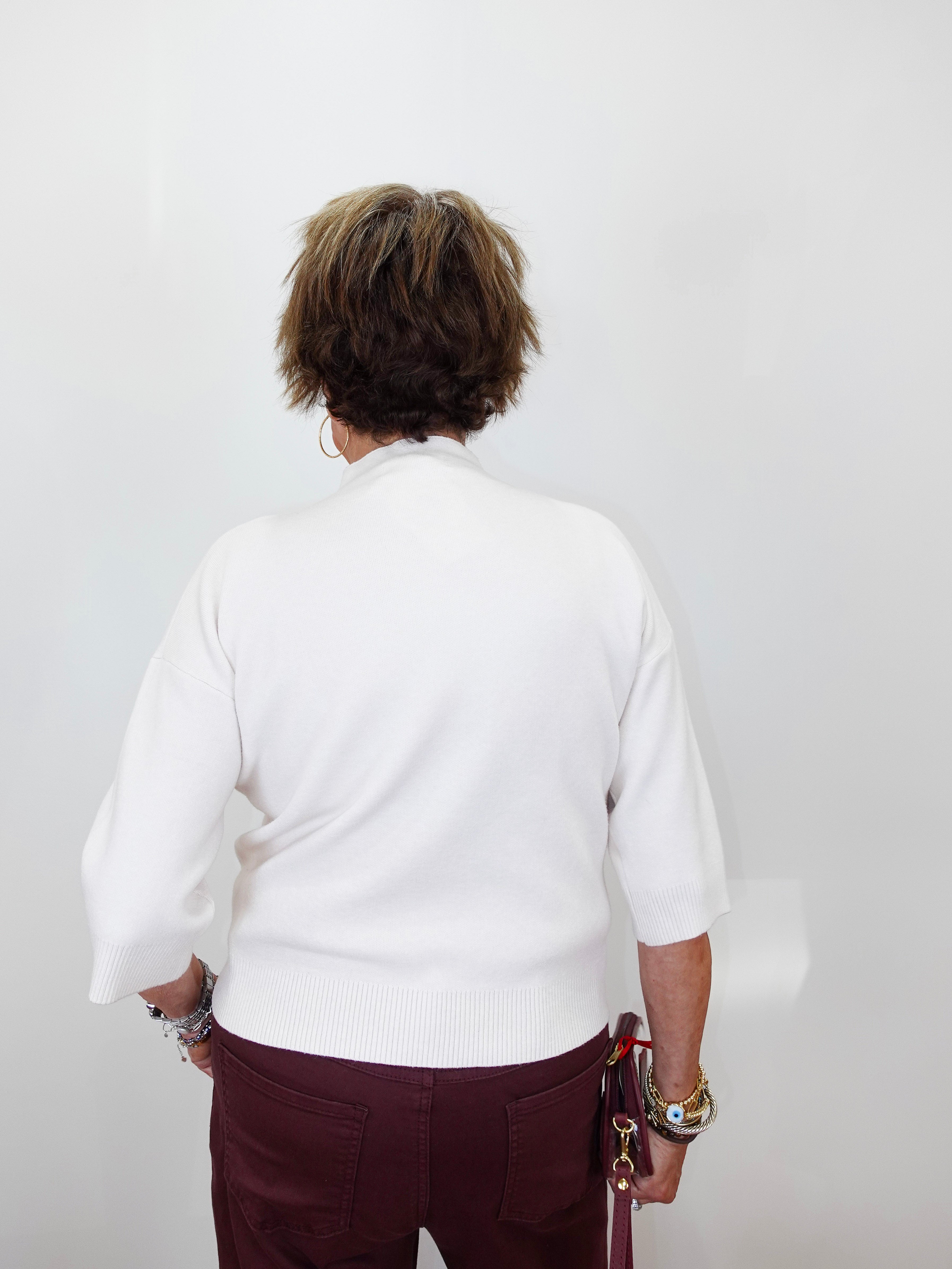 A person viewed from behind wearing a white knit sweater with three-quarter sleeves and burgundy pants, standing against a plain light gray wall.