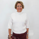 A woman with short blonde hair wearing a white mock-neck knit top and burgundy pants, holding a metallic burgundy handbag against a plain white background.