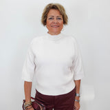 A woman with short blonde hair wearing a white mock-neck knit top and burgundy pants, holding a metallic burgundy handbag against a plain white background.