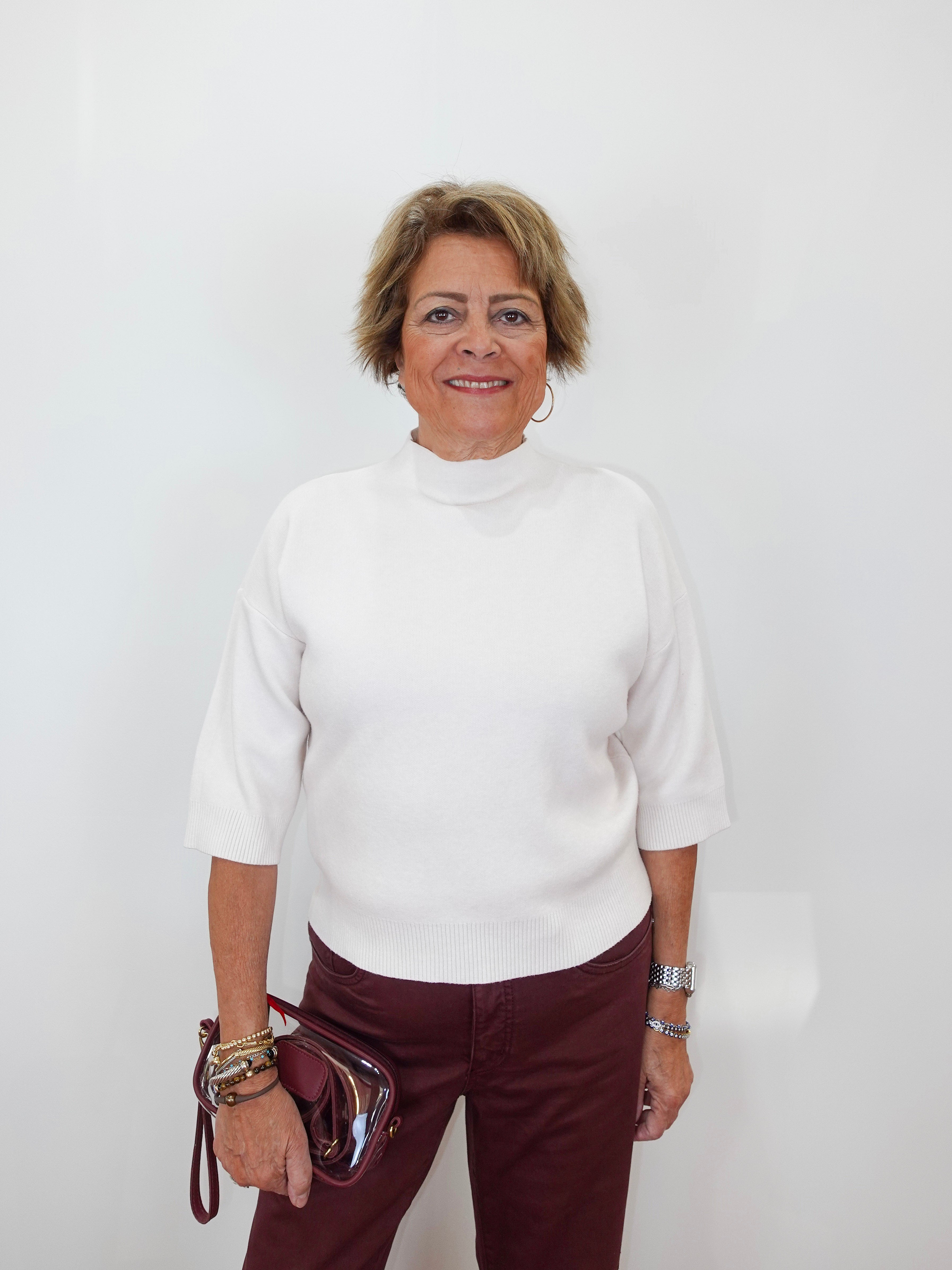 A woman with short blonde hair wearing a white mock-neck knit top and burgundy pants, holding a metallic burgundy handbag against a plain white background.