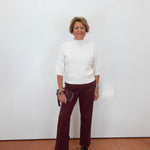 A woman stands against a plain white wall wearing a white mock-neck knit top, burgundy wide-leg pants, and black sandals, holding a small dark clutch.