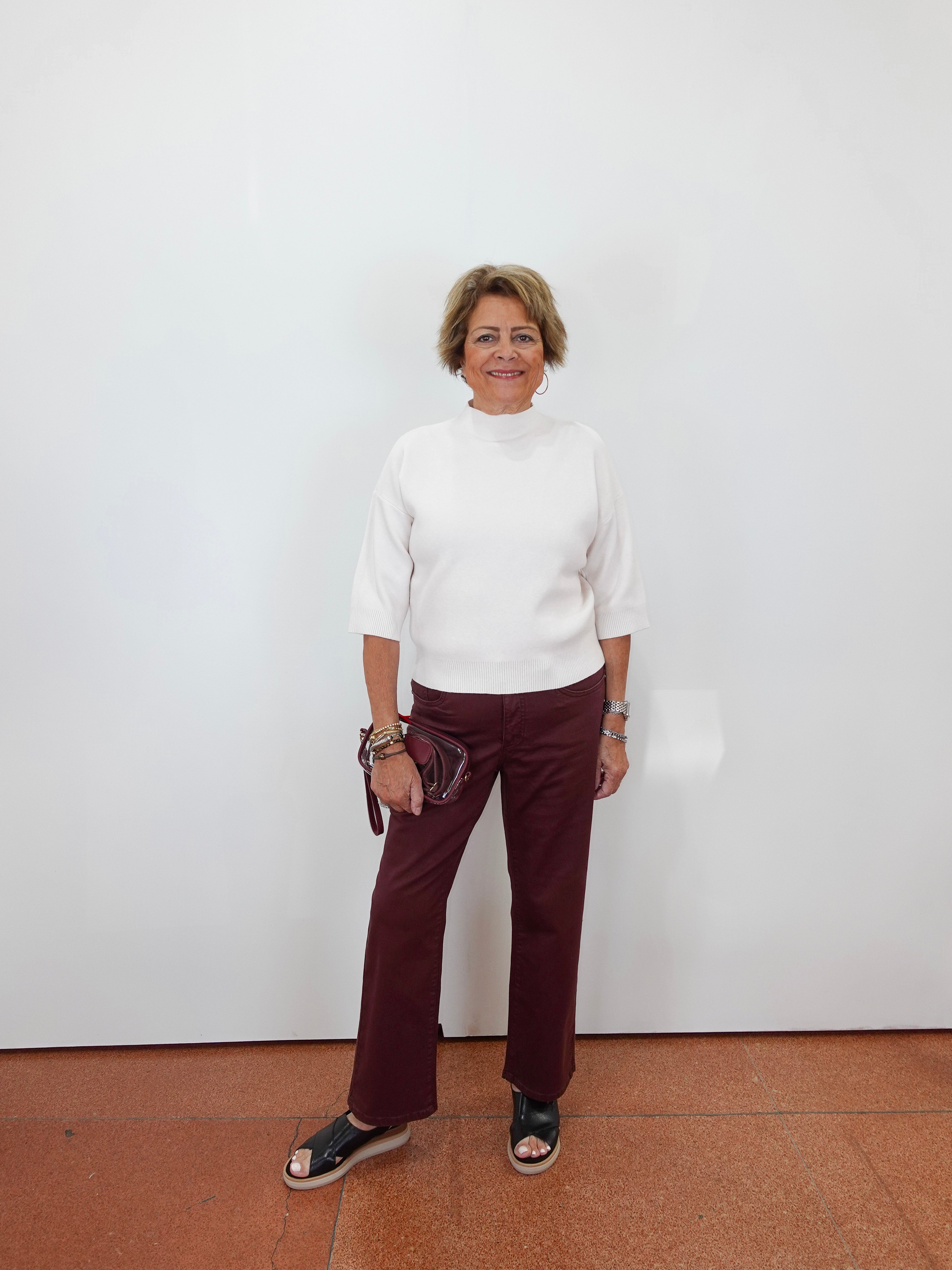 A woman stands against a plain white wall wearing a white mock-neck knit top, burgundy wide-leg pants, and black sandals, holding a small dark clutch.