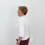 An older woman with short brown hair is shown in profile against a plain white wall, wearing a white three-quarter-sleeve top and dark burgundy pants.